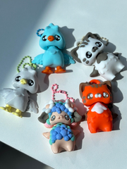 Multiple 3D printed sheep bag charms with pastel rainbow colors and white flower details on ears, Orange Cat,White cat ,blue goose and white goose scattered on the floor