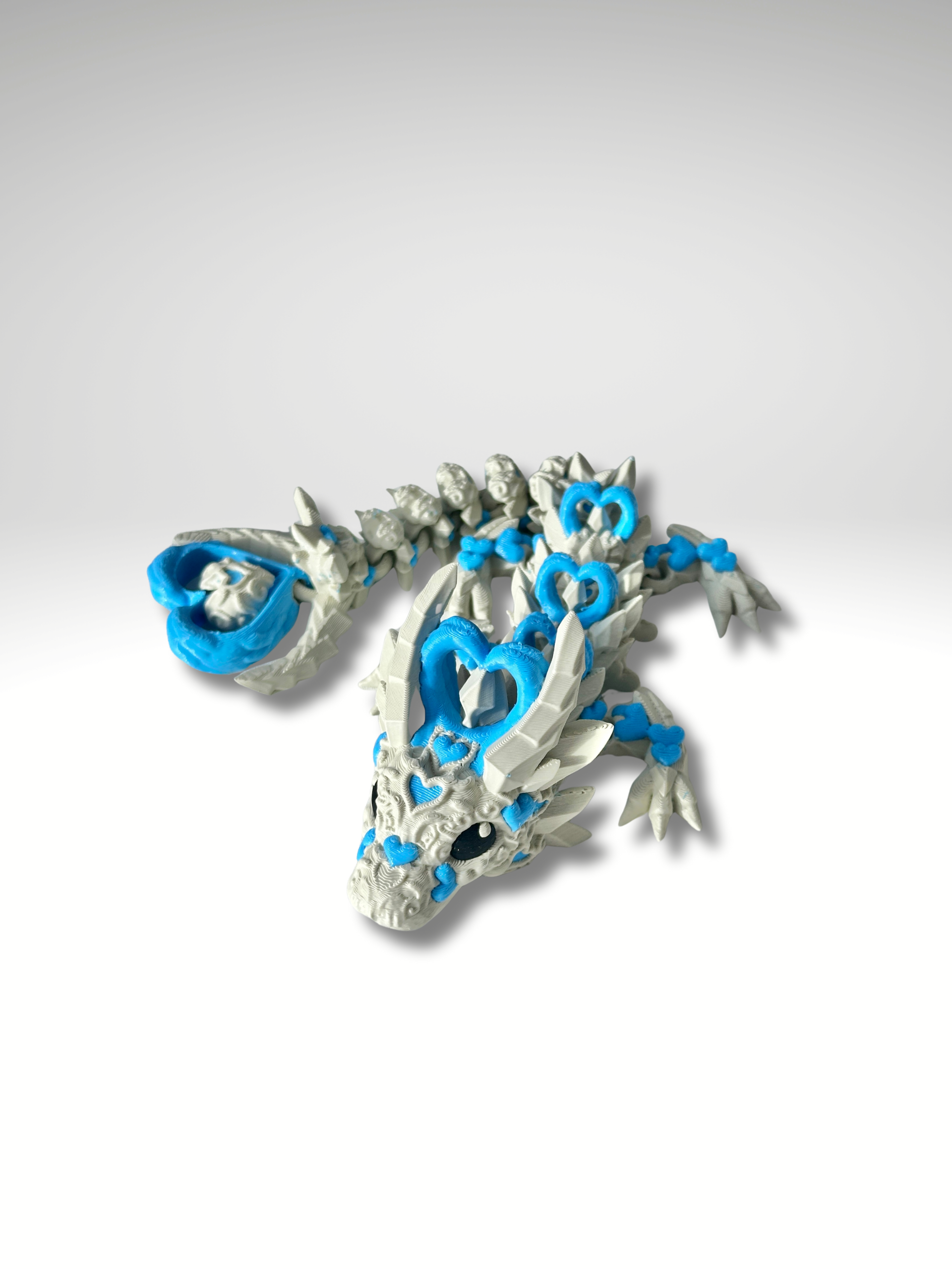 3D printed articulated Heart Dragon in Khaki-Blue posed in a curled position, showing front view of head, body segments, and heart-tail