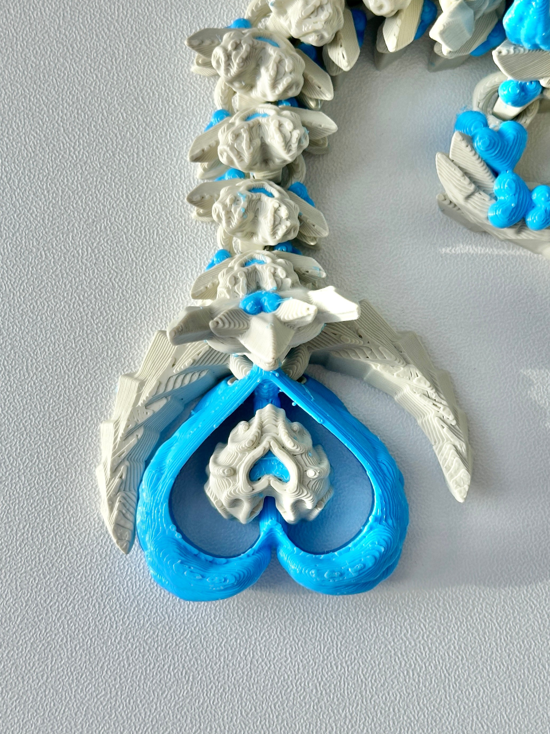 Close-up of the Heart Dragon’s tail showing a blue heart-shaped tip with a small beaded heart center, surrounded by feather-like textures