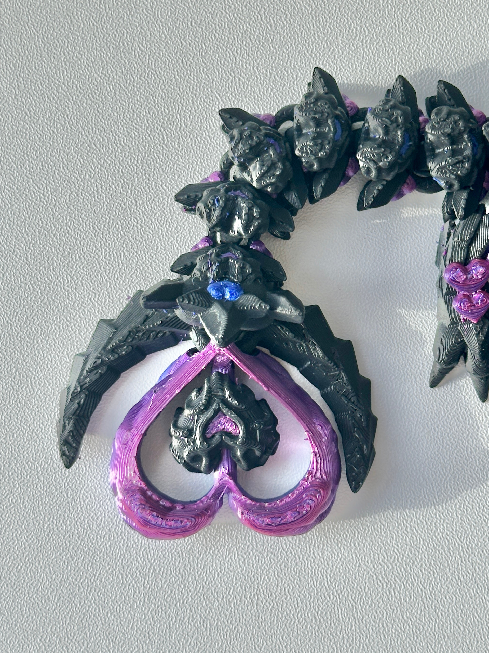 Close-up of the Heart Dragon’s tail showing a purple heart-shaped tip with a small beaded heart center, surrounded by feather-like textures